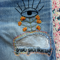 Embroidered eye and flowers design on denim fabric with 'grow your own way' text.