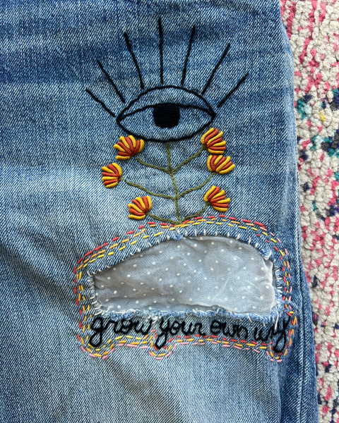 Embroidered eye and flowers design on denim fabric with 'grow your own way' text.